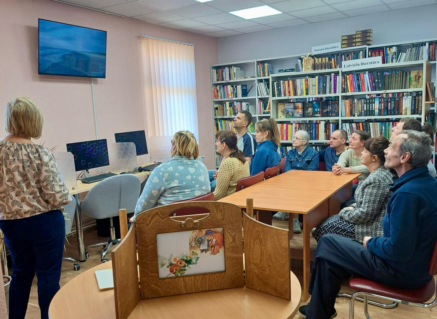 Cilvēki bibliotēkā skatās video ekrānā
