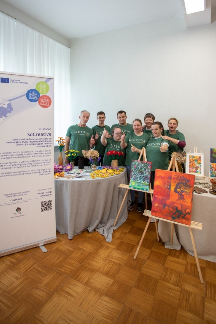 Cilvēku grupa stāv pie galda, priekšplānā košas gleznas