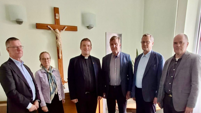 Cilvēku grupa stāv puslokā pie koka krusta