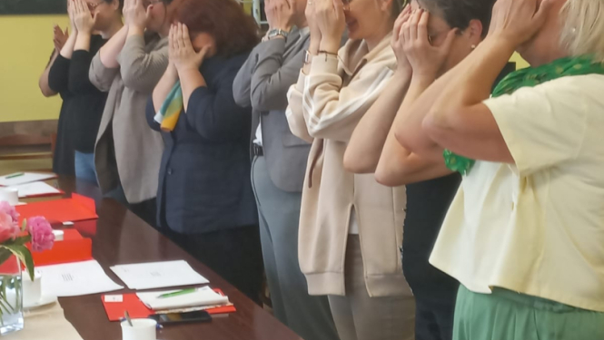 Cilvēki stāv ap galdu, ar rokām aizklājuši sejas