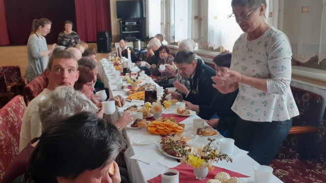 Cilvēki sēž pie svētku galda ar  ēdieniem, stāvoša sieviete viņus uzrunā