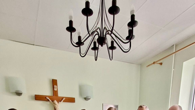 Cilvēku grupa stāv puslokā pie koka krusta