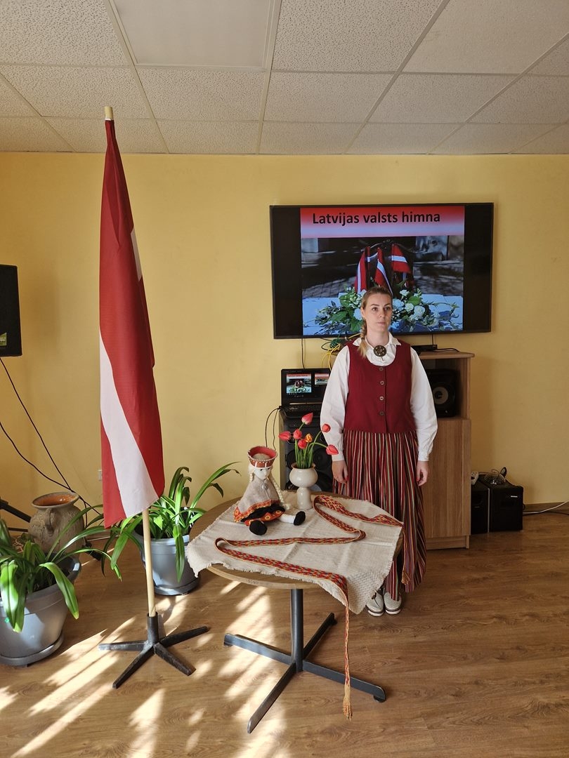Sieviete tautastērpā stāv pie Latvijas kroga, uz galda vāze ar ziediem