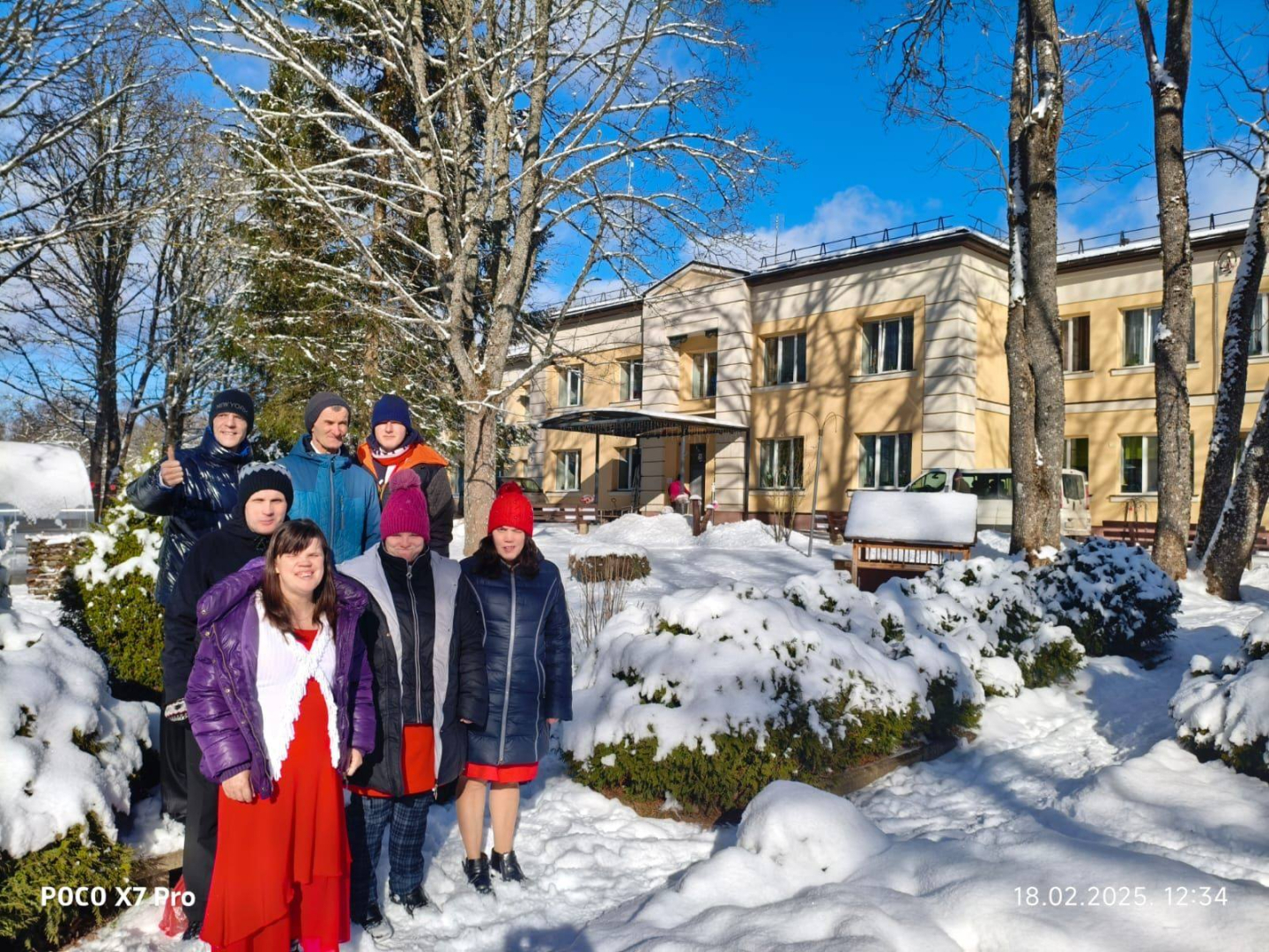 Cilvēku grupa ārā ziemas dienā stāv pie apsnigušiem apstādījumiem