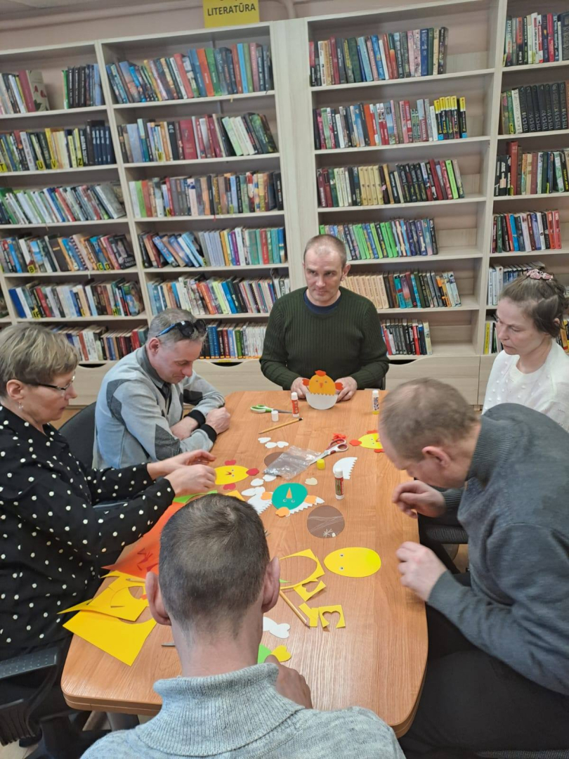 Cilvēki ap galdu veido rokdarbus bibliotēkā