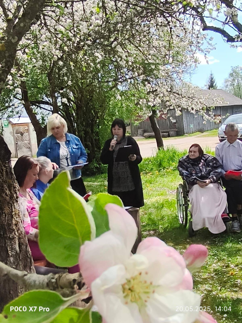 Cilvēku grupa stāv zem ziedošu ābeļu zariem