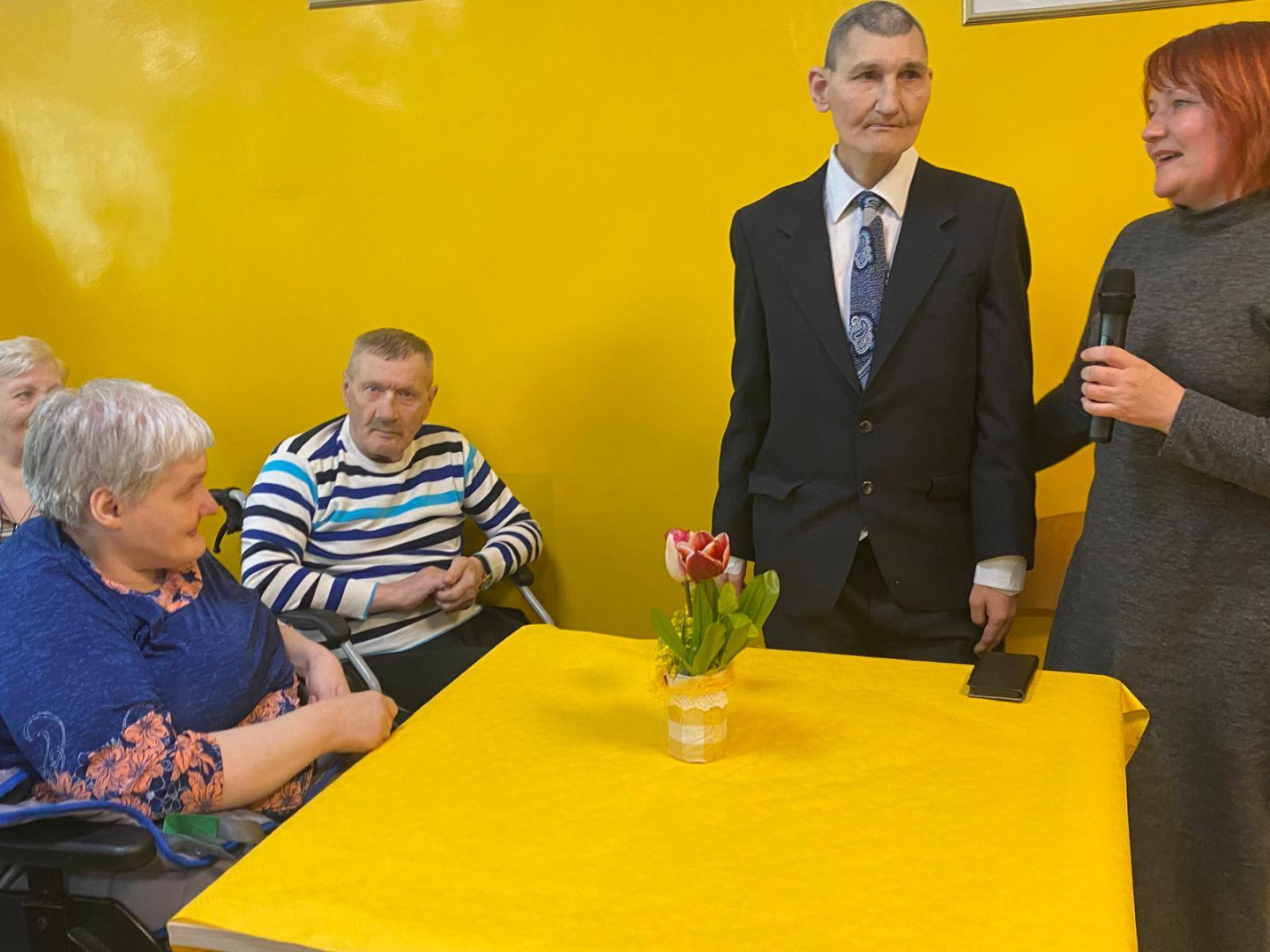 Sieviete pie dzeeltena galda, pie galda stāv sieviete un vīrietis