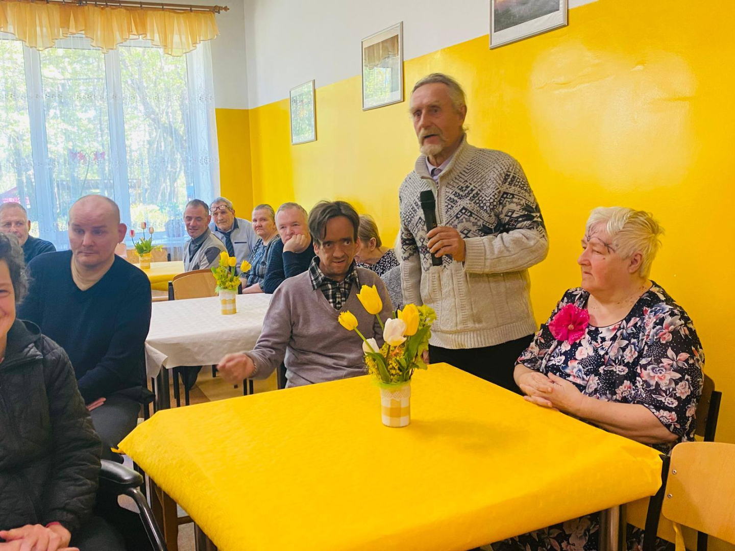 Cilvēki ap dzeltenu galdu, vīrietis stāv un runā mikrofonā