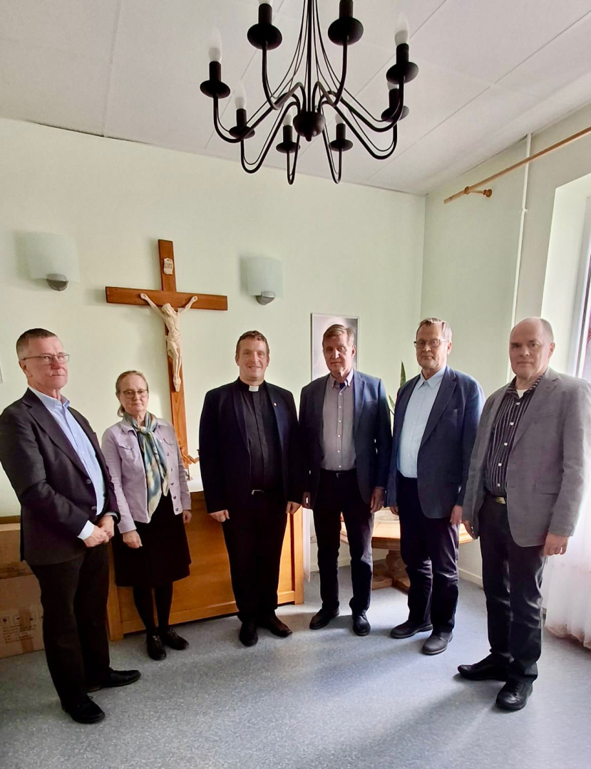 Cilvēku grupa stāv puslokā pie koka krusta