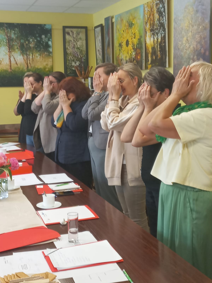 Cilvēki stāv ap galdu, ar rokām aizklājuši sejas