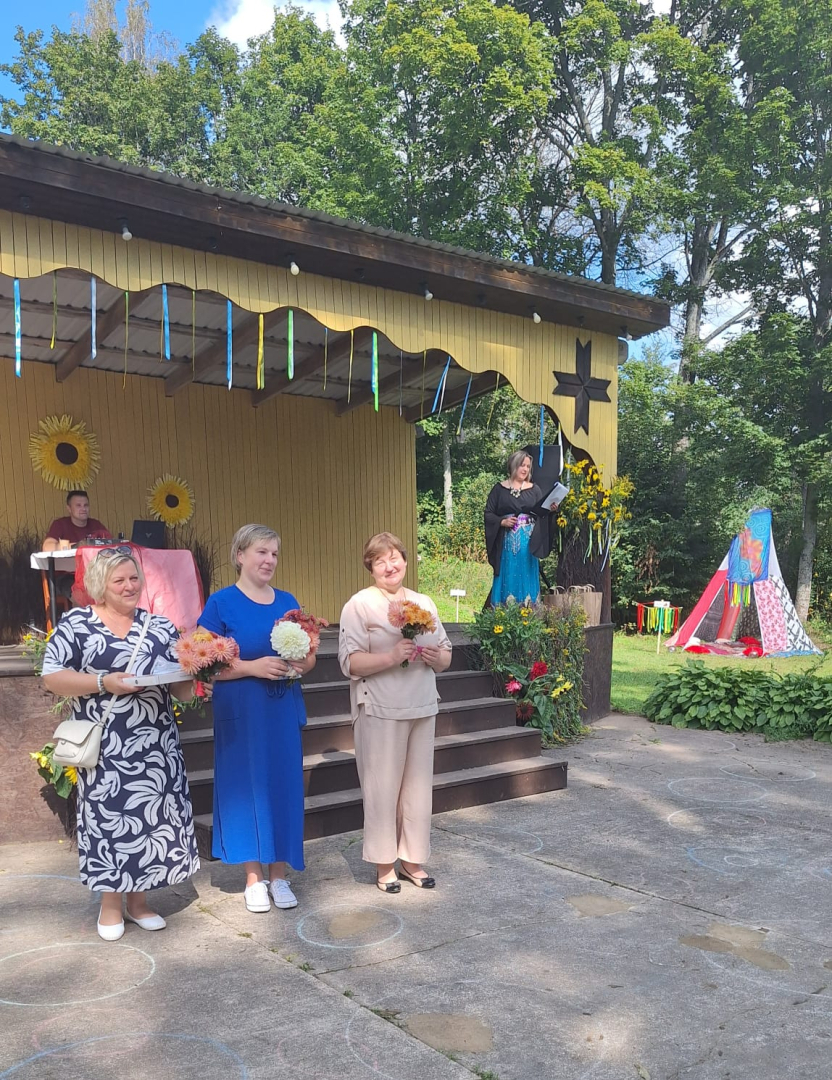 3 sievietes uz skatuves, ar ziediem rokās, saka uzrunu