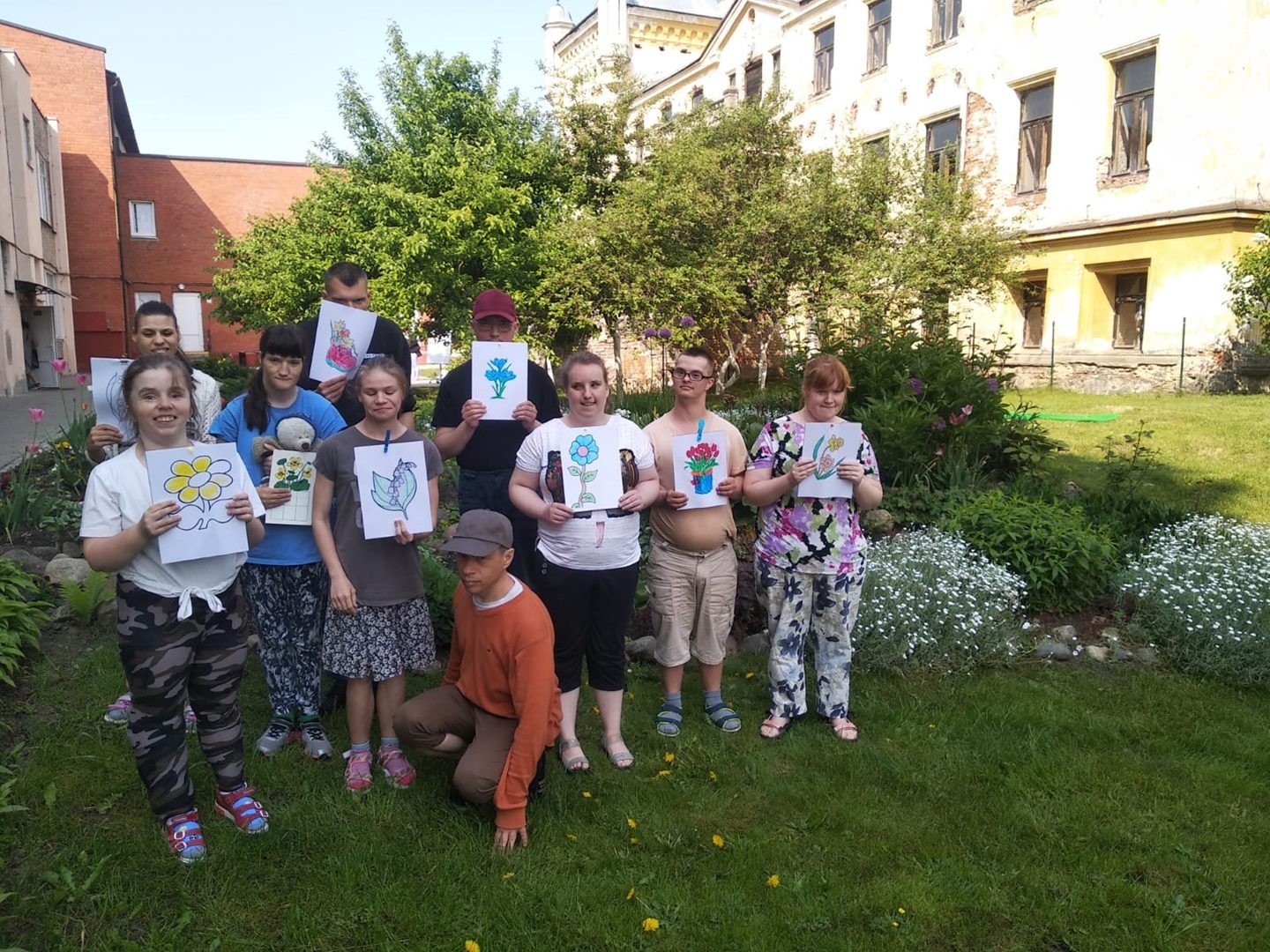 Cilvēku grupa stāv zālājā, rokā ziedu zīmējumi