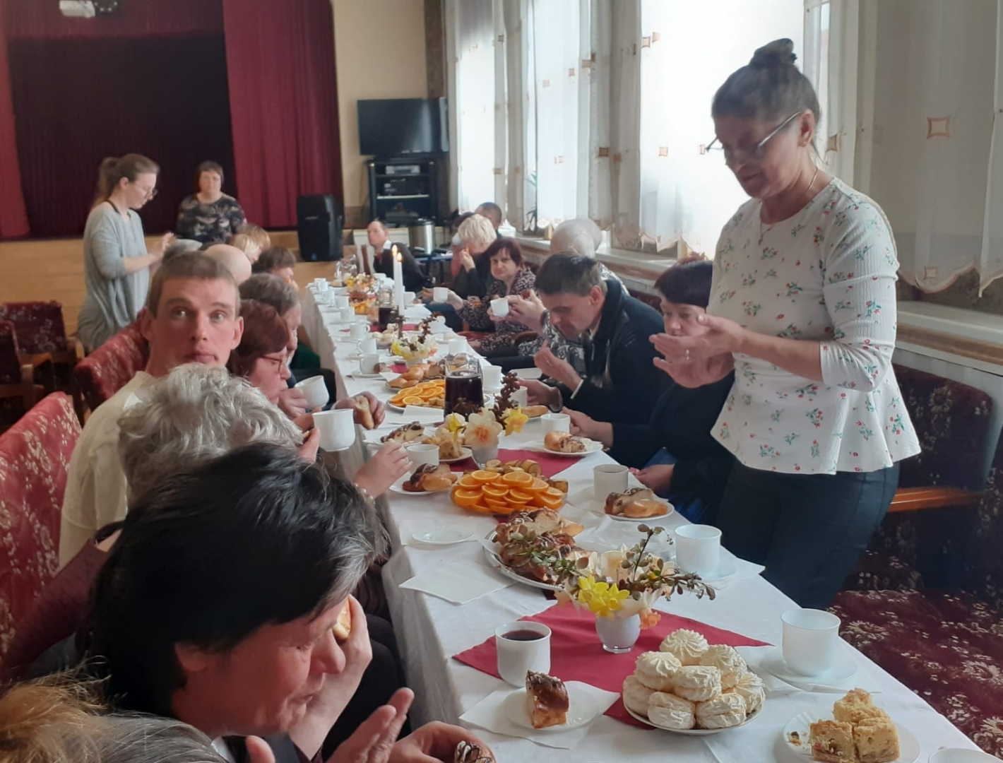 Cilvēki sēž pie svētku galda ar  ēdieniem, stāvoša sieviete viņus uzrunā