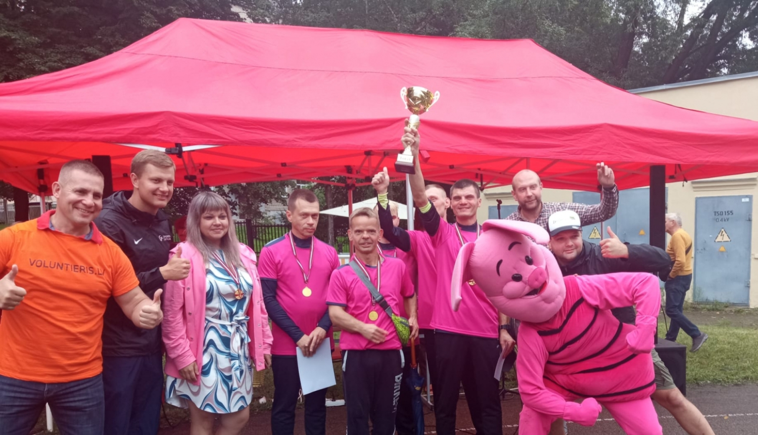 Sportisti stāv pie telts un tura kausu