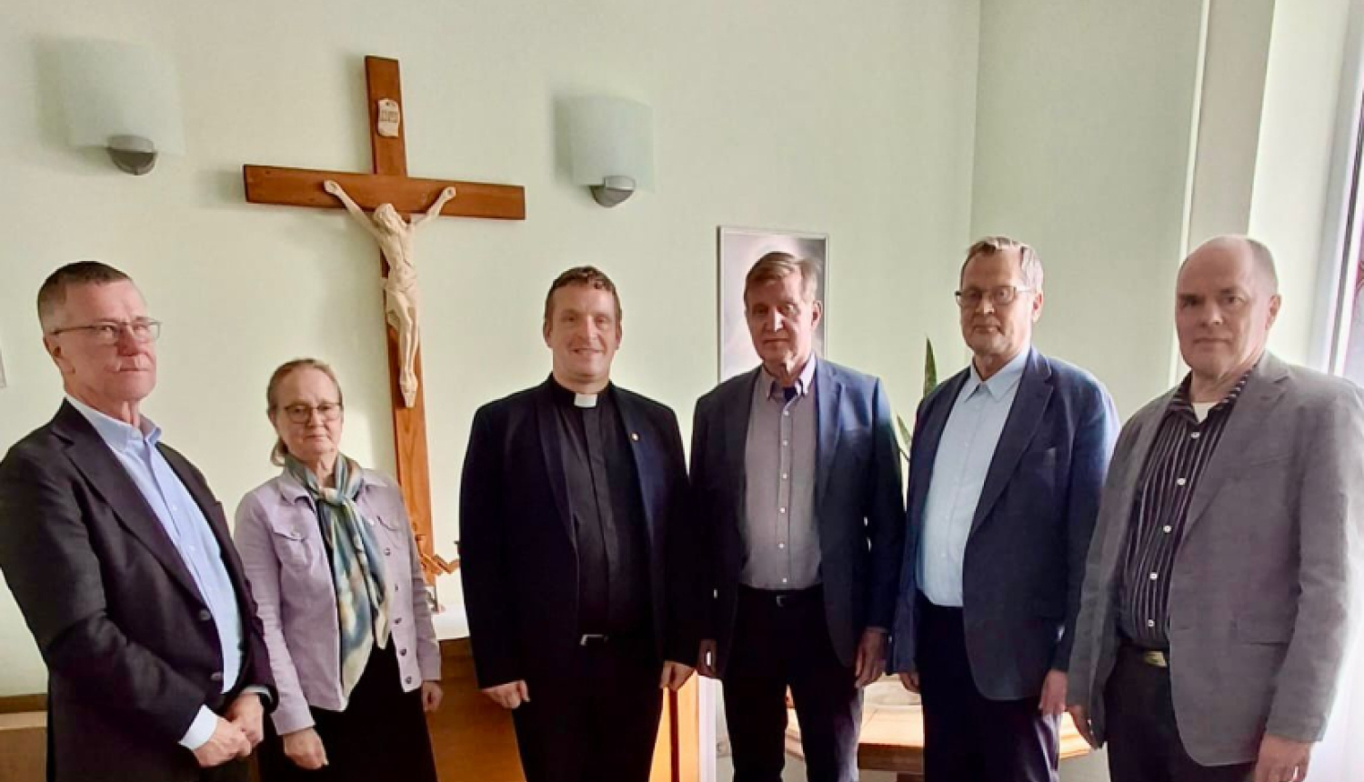 Cilvēku grupa stāv puslokā pie koka krusta